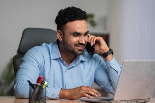 hombre de negocios indio hablando por teléfono móvil mientras trabaja en una computadora portátil en la oficina: concepto de asesor financiero, multitarea y estilo de vida corporativo - multi tasking efficiency financial advisor business fotografías e imágenes de stock