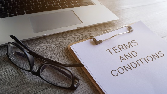 Paper clipboard with text TERMS AND CONDITIONS on table with laptop and glasses.