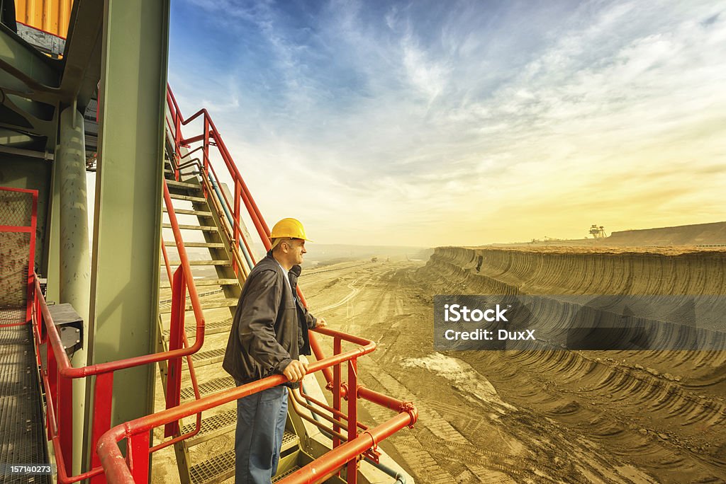 Happy worker on drill machine Smiling and happy electrical engineer standing and looking at the side. In the background is a huge coal digging site with beautiful cloudy and colorful sky. Photographed with wide angle lens. Mining - Natural Resources Stock Photo