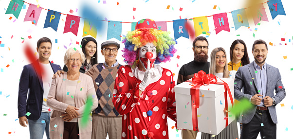 Clown holding a surprise present at a birthday party with people gathered behind isolated on white background