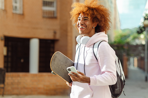 Portrait of a cheerful African American man with wireless headphones using a smart phone and carrying his skateboard while walking