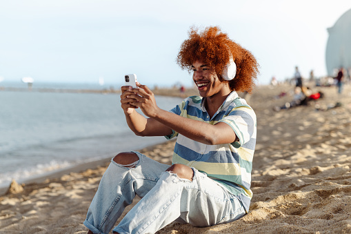 Portrait of a smiling African American man taking a selfie with his smart phone while having a relaxing time on the beach