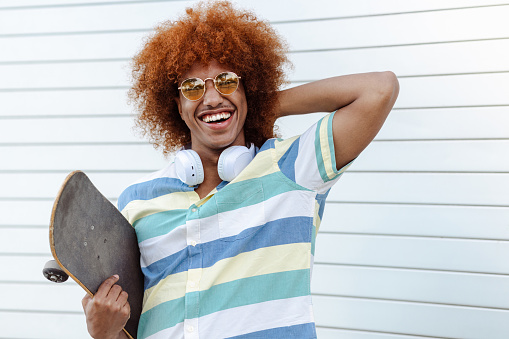 A Black man with a cool attitude is standing against a white background and looking at the camera. He is holding a skateboard. Space for copy.