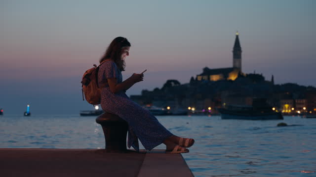 SLO MO Woman with Backpack Using Smartphone on Harbor with Old Town of Rovinj at Night