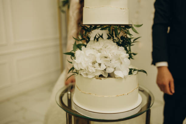 istock Three-tiered white wedding cake decorated with flowers from mastic on a white wooden table. Picture for a menu or a confectionery catalog with copy space. 1571530219