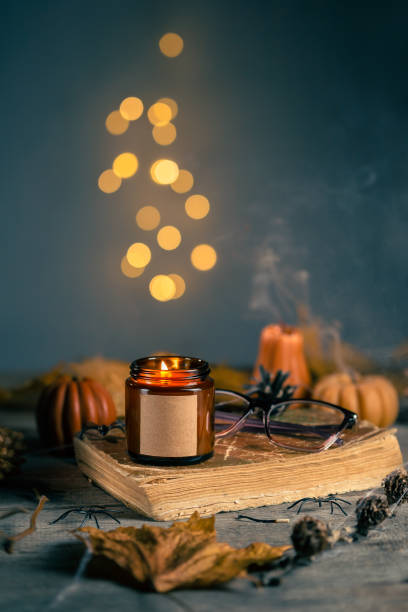Halloween Still Life with Soy Wax Candle in the Dark Amber Glass Jar with Empty Label for Mock Up. Pumpkin Shaped Candles with Cones, Leaves, Old Book and Glasses. Bokeh Lights on the Background