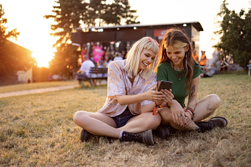 Happy female friends surfing the Internet on cell phone while relaxing on grass during a music festival at sunset.