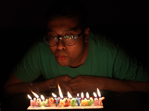 A South Asian man in glasses, blowing out the candles on a birthday cake in the darkness