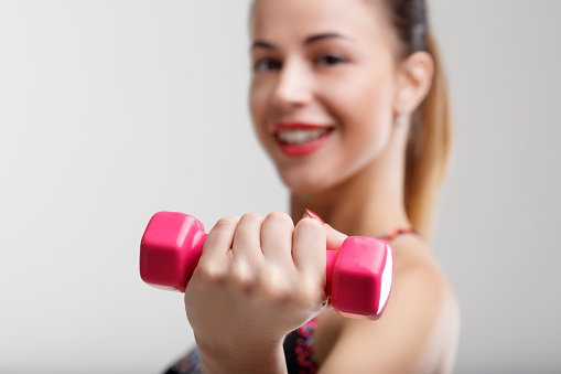 In-focus small pink weights with a blurred woman in the background. She enjoys her light fitness workout which adds happiness and positivity to her day
