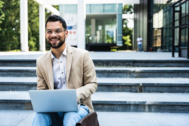 istock Handsome businessman in glasses working on his laptop computer while resting in city centre. Freelance man typing something in his document. Office worker finishing work outside of office building. 1571671135