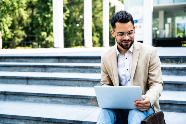 istock Handsome businessman in glasses working on his laptop computer while resting in city centre. Freelance man typing something in his document. Office worker finishing work outside of office building. 1571671165
