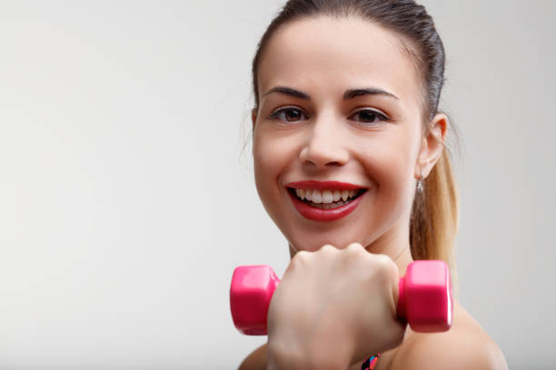 Foreground shows blurred weight, background features a woman working out. Her light exercise with small pink weights brings pride and mental uplift. A smiling start to the day with tied-up hair