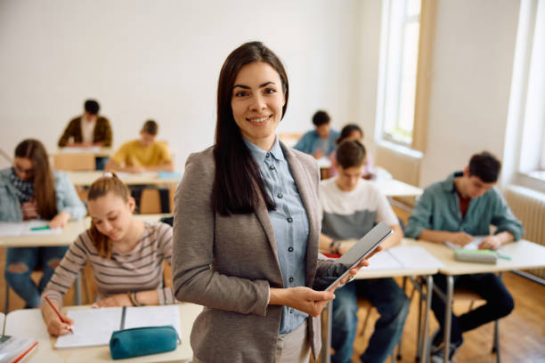 istock Smiling high school teacher in the classroom looking at camera. 1571674422