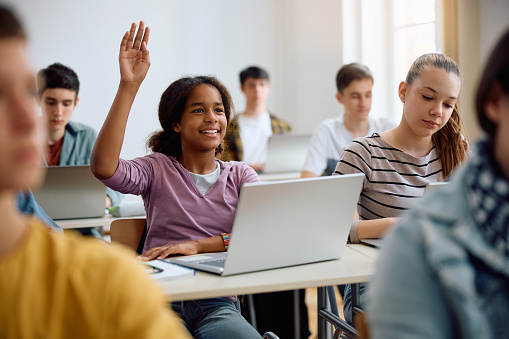 Happy African American teenage girl using laptop and raising her hand to ask a question during computer class at high school.