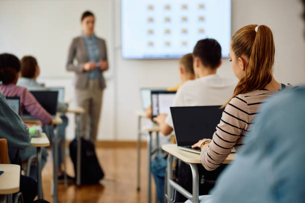 istock Back view of teenage girl using laptop during computer class at high school. 1571674952
