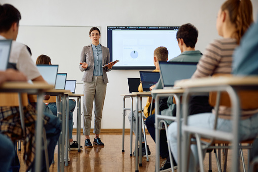 Teacher With College Students Giving Lesson In Classroom Holding A Digital Tablet