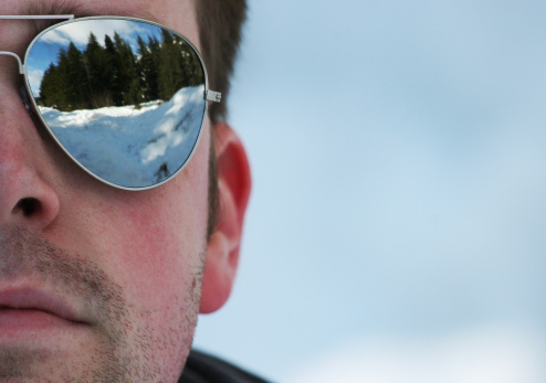 Half of a man's face wearing reflective sunglasses with a stubble beard. Reflection shows trees and snow.