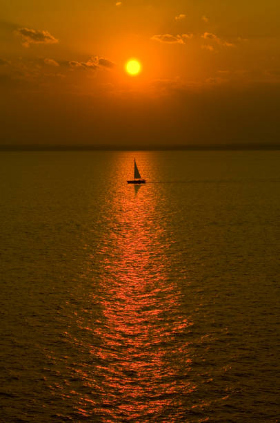 A sail boat out at sunset, vertical shot with sailboat lined up with the sun and its reflection on the water.