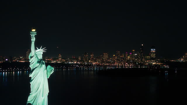 Cinematic Helicopter Pass by the Statue of Liberty at Night. Close Up Panoramic Aerial Footage of the Illuminated Roman Goddess Holding a Torch. Landmark New York Monument with Manhattan Skyscrapers