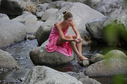 Portrait of beautiful young woman in bright pink dress posing on the large stones in the riverbed. Concept of wellbeing, mindfulness, connecting with nature and enjoying the healing sound of water