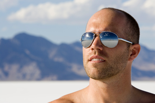 An attractive guy stands alone in the middle of the Salt Flats in Utah with the scene of emptiness reflected in his sunglasses.