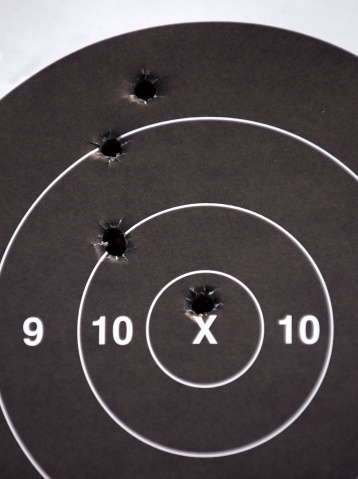 A paper target marked with real bullet holes, one clearly in the bullseye ring.