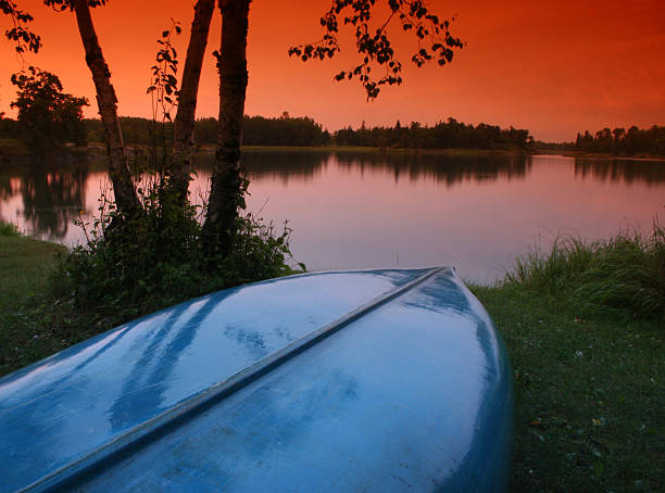 A canoe sits on the shore of a pristine lake. Blue canoe and pristine muskoka lake in Ontario, Canada. Beautiful sunset over the calm water of the freshwater lake. Themes include canoeing, paddling, recreation, leisure, lake, lakefront, cottage, muskoka, Ontario, Canada, shore, wet, sunset, scenic, landscape, beauty, nature, and Algonquin park. Nobody is in the image. 
