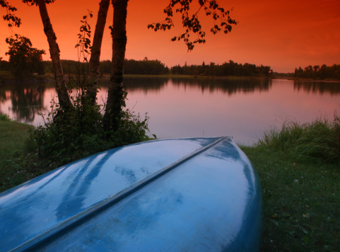 A canoe sits on the shore of a pristine lake. Blue canoe and pristine muskoka lake in Ontario, Canada. Beautiful sunset over the calm water of the freshwater lake. Themes include canoeing, paddling, recreation, leisure, lake, lakefront, cottage, muskoka, Ontario, Canada, shore, wet, sunset, scenic, landscape, beauty, nature, and Algonquin park. Nobody is in the image. 