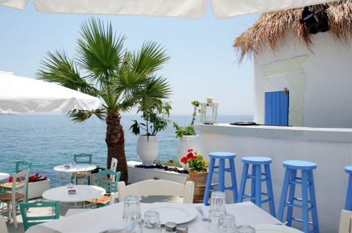 View of an idyllic, open-air, outdoor restaurant in Greece overlooking the Aegean sea. White walls, blue decoration, thatched roof and a palm tree, with the Aegian sea in the background. The perfect place for a relaxed lunch on a hot sunny, summer day in Greece.