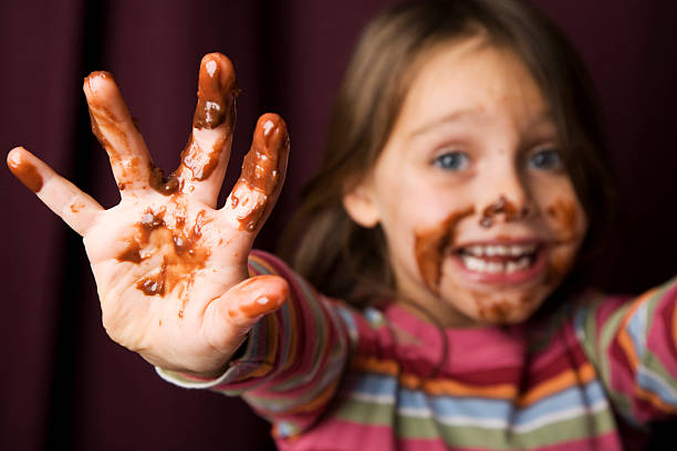 Young Girl Covered in Chocolate. Young girl with chocolate smeared all over her face and fingers. Focus is on mucky hand with face out of focus against a rich red background. chocolate fingers stock pictures, royalty-free photos & images