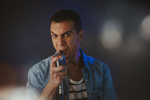 A retro male rock singer in a jeans shirt is holding a vintage microphone with stand and sings.