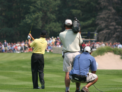 A camera crew covers a golf tournament. Media coverage of major sporting event with camera man and cable man or sound man behind. Golfer is making a swing with gallery of golf fans behind the green. Additional themes include media, coverage, filming, crew, sound, technical, taping, video, sports, event, recording, documentary, movie, broadcasting, sports coverage, and major sporting events. 