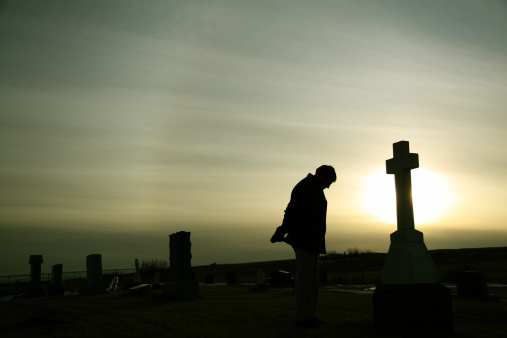 A man remembers a loved one beside a tombstone at a graveyard. Additional themes include death, loss, funeral, remembering, tribute, memorial, memorial day, veteran's day, sadness, heaven, cemetery, and grief. Unrecognizable man in 30s. 