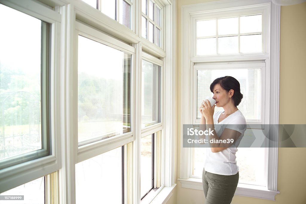 woman standing by a window drinking coffee A woman sips coffee by a window Adult Stock Photo