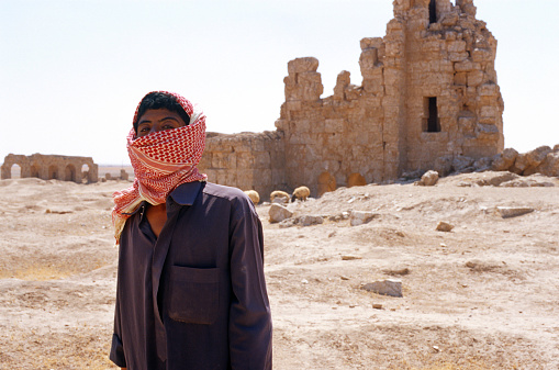 Young Arabic shepherd in traditional Kaffiyeh and Jellabiya, and his sheep on a desert among ruins background, Syria.