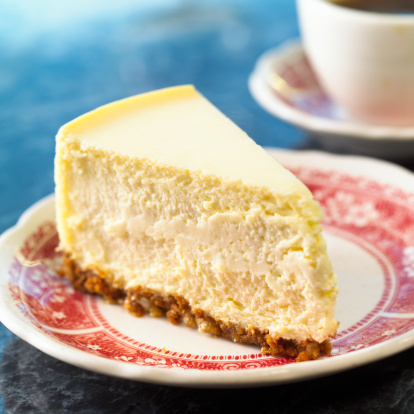 Plain Cheesecake on red plate with coffee in background.  Shot with shallow focus on front of cake.