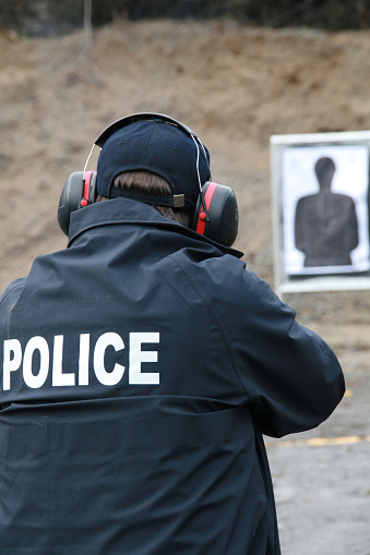Picture of a Policeman shooting handgun in a practice field.  The police officer is wearing his uniform and has earmuffs to protect him from the sound of the handgun. The shooting target is blur and visible in the background. 