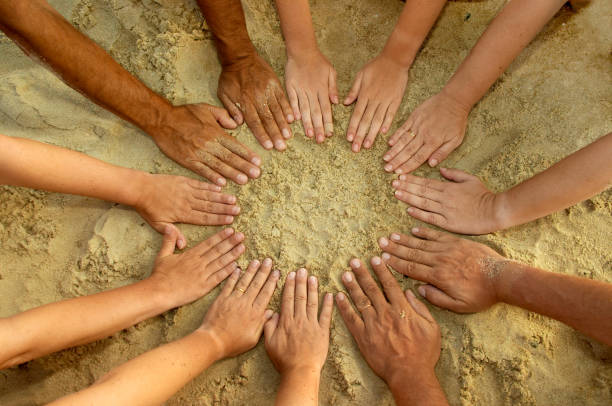 High angle view of a circle of hands touching sand. Horizontal format.
