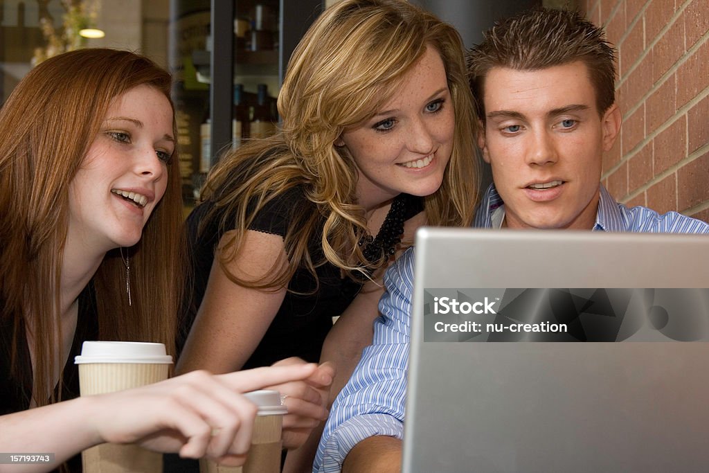 Group Meeting 2 young women and one young man at a coffee shop looking at a laptop screen and pointing. High School Stock Photo