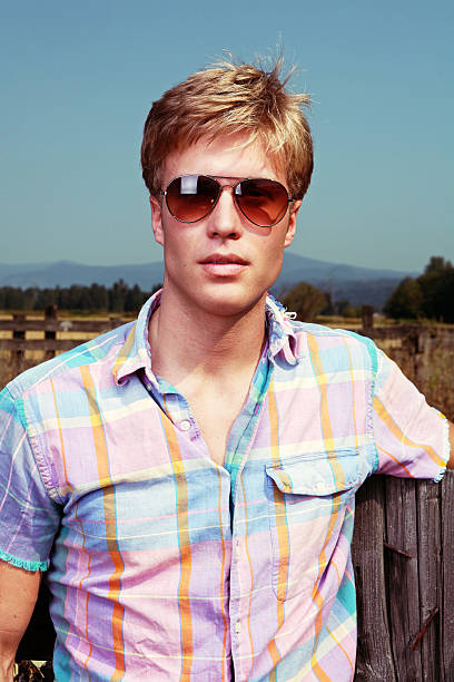 A man in a standing in front of a field wearing sunglasses and a shoulder-bag. 