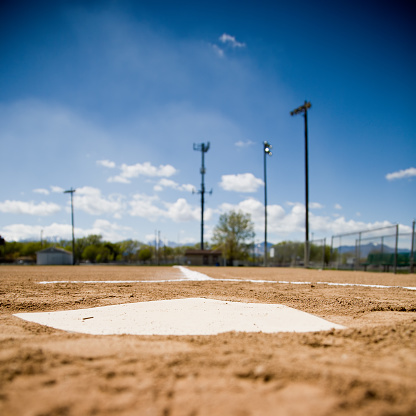 A close up of home plate on a baseball diamond.  Shot from a very low angle.  The blue sky and the lights are visible in the out of focus background.
