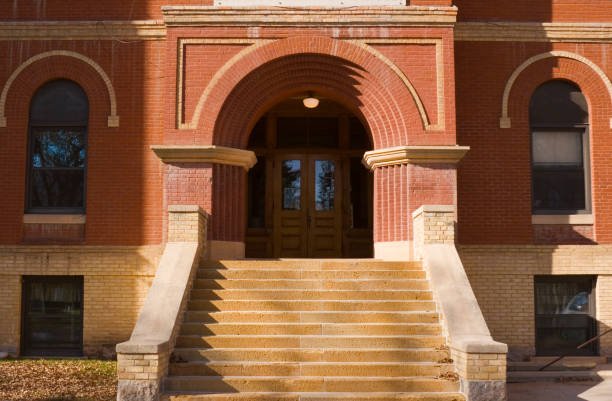 Horizontal straight-on exterior view of an old red brick high school or elementary schoolhouse building facade, centered on the entrance front door and staircase.  The traditional architecture suggests a respect for higher education and learning. The brownstone built structure is located in a small Midwestern town in the U.S.A.