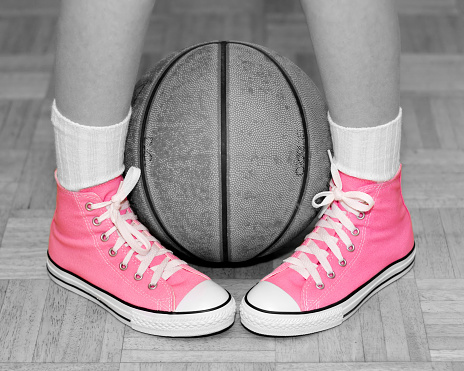 A shot of a girl's feet wearing pink basketball shoes with a basketball.  Only the shoes have color, everything else is black & white.