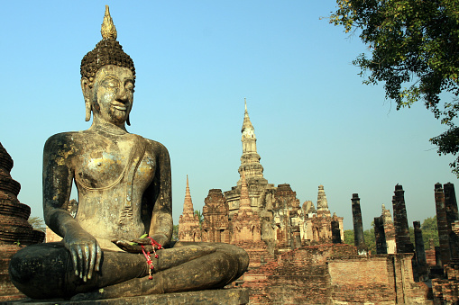 DSLR picture of a Buddha at sunrise in the famous temple of Sukhothai in Thailand. The buddha is in the foreground of the picture ans there is a temple in the background. The sky is blue without cloud. 