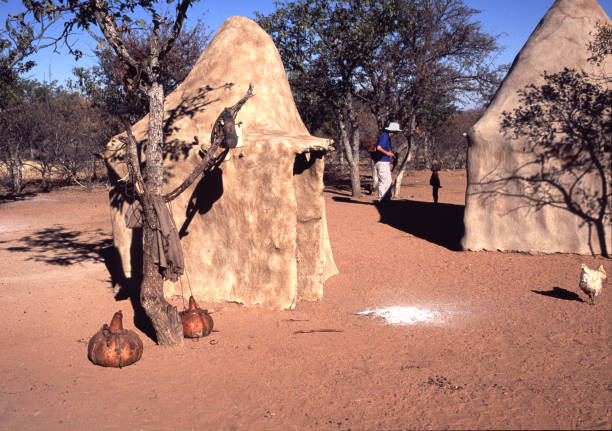 The Himba are the only traditionally-living tribe in Namibia. They live in a very hot, sunny, arid area. This photo shows part of a village ij Kakaoland, with huts, gourds for churning butter and ashes from the sacred fire in front of the headman's hut. A tourist is meeting a child from the village.