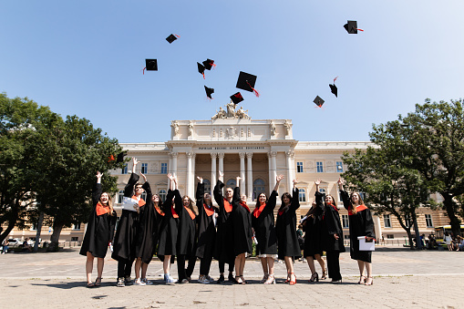 In this joyous and celebratory scene, a group of cheerful students, dressed in their graduation robes, enthusiastically throws up their square academic caps into the air as a symbol of their university graduation. With wide smiles and jubilant expressions, they capture the exhilaration and sense of accomplishment that comes with completing their academic journey.