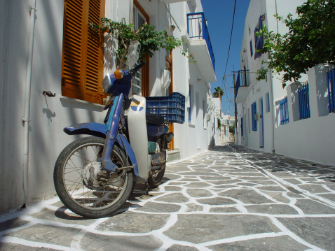 Low angle view of a blue scooter parked  in a traditional Greek street on one of the Cyclades Islands in the Cyclades Sea. The street is lined with white houses and the street is partly painted white. Both to protect against the heat during summer.