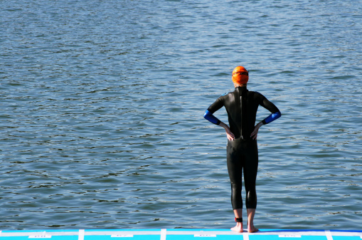 A lone female swimmer in black wetsuit and orange cap standing at the edge of the water contemplating the start of a triathlon race.