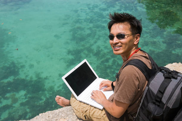 Asian male hiker taking a break to check email at the edge of a cliff looking down into the water.
