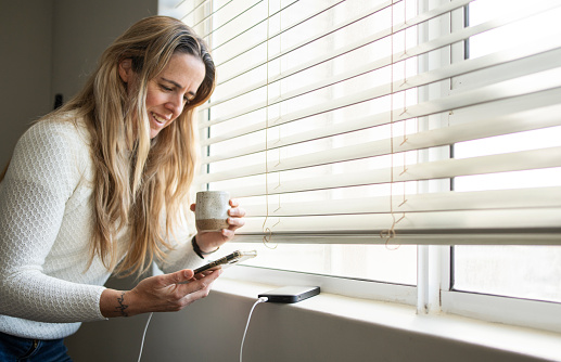 Smiling woman drinking coffee by a window at home and reading a text message on her phone charging with a power bank